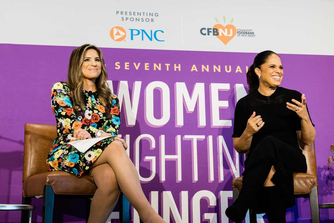 Soledad O'Brien and Natalie Pasquarella conversing on a stage in front of a Women Fighting Hunger Luncheon banner